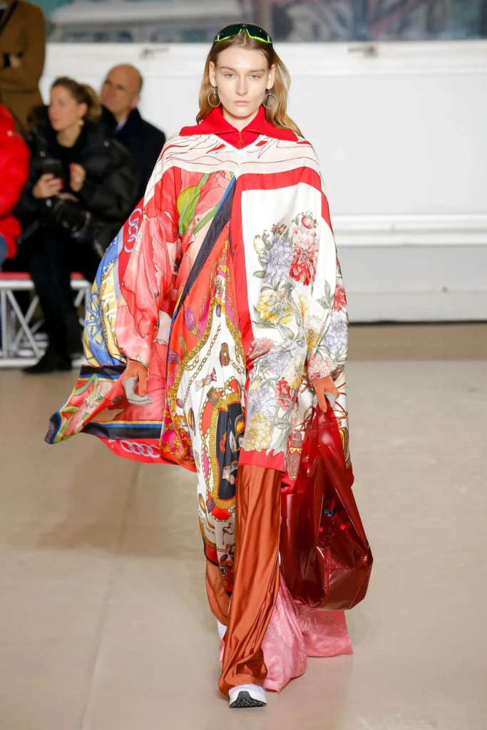 Une mannequin au défilé de mode de Marine Serre avec une tenue d'upcycling à partir de tissus très légers qui forment un manteau coloré rouge et blanc avec des motifs.